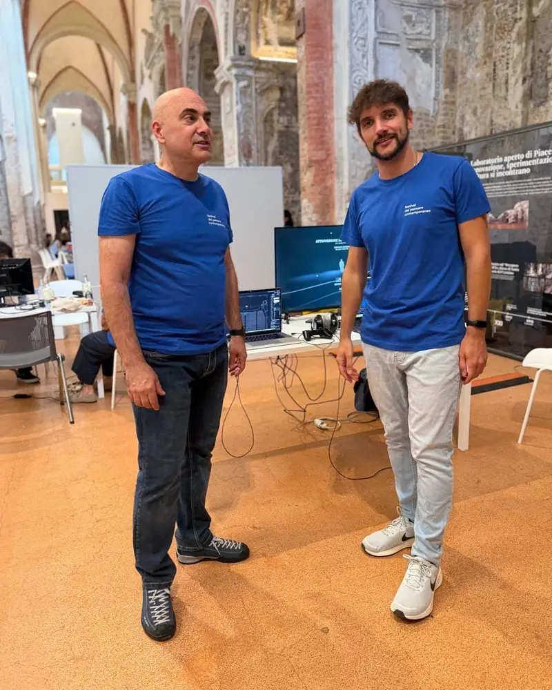 Two people in the festival’s blue T-shirt standing in the nave of the Carmine; behind them the station and a screen reading “Attraversare il suono”.