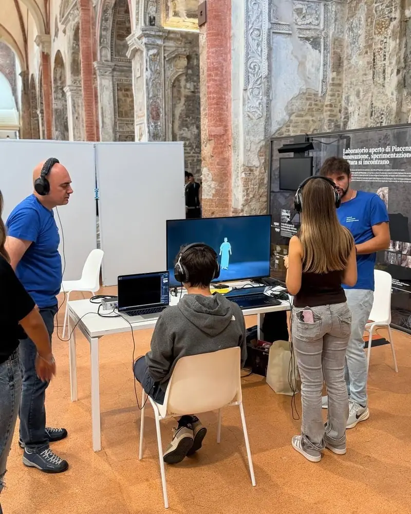 Under the frescoed arches of the Carmine, a person seated with their back to the camera, wearing headphones, looks at a screen with Calaf on stage; beside them another person standing with their back turned and two people in the festival’s blue T-shirt.